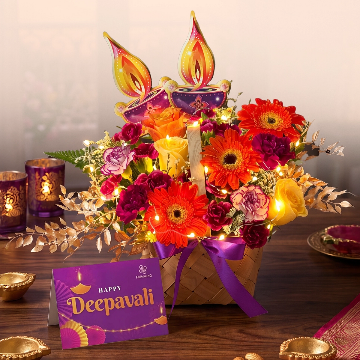 A festive Diwali flower arrangement in a woven basket with purple ribbon, decorated with orange and red gerbera daisies, pink and yellow roses, gold leaves, and two lit diya candles. A purple 'Happy Deepavali' card sits on the table beside purple lanterns.