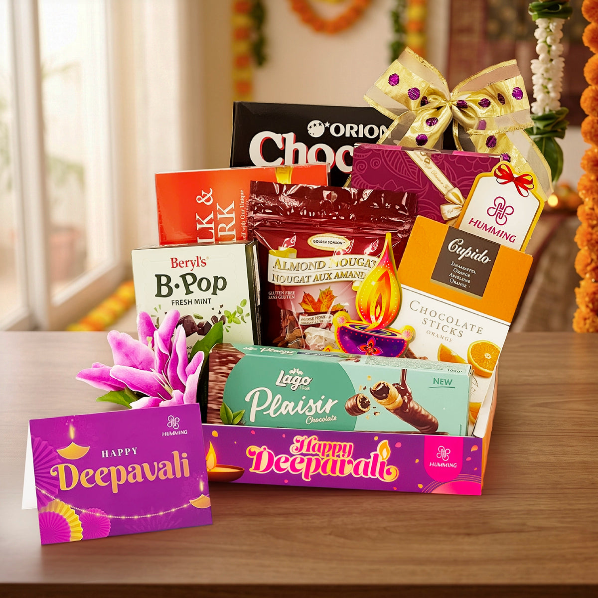 A festive Deepavali gift basket filled with assorted chocolates, sweets, and packaged treats, arranged on a wooden table with a decorative bow and a purple Happy Deepavali card in front.