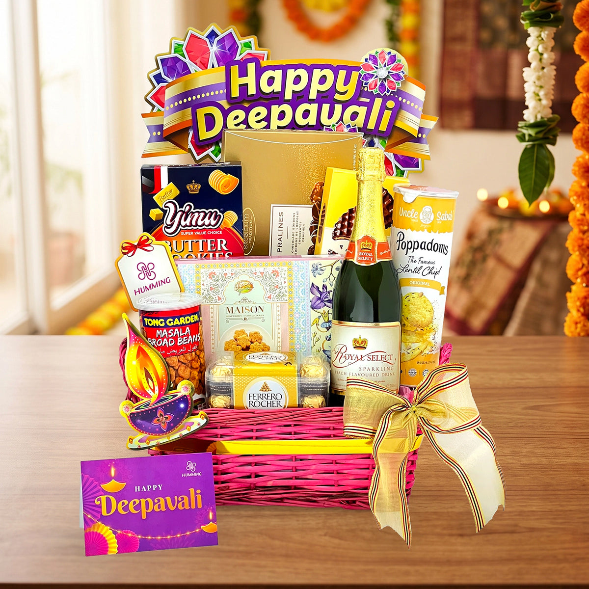 A festive Deepavali gift basket filled with assorted snacks, chocolates, and a bottle of sparkling wine, arranged on a wooden table with a pink wicker basket, gold ribbon, and a purple 'Happy Deepavali' card.