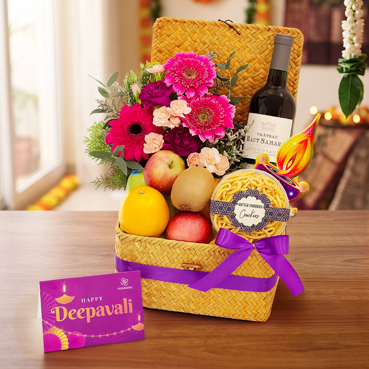 A festive Deepavali gift basket arranged on a wooden table, featuring a woven yellow basket with a purple ribbon, a bottle of wine, a bouquet of bright pink and red flowers, apples, a lemon, a packaged snack, and a pink 'Happy Deepavali' card placed beside it.