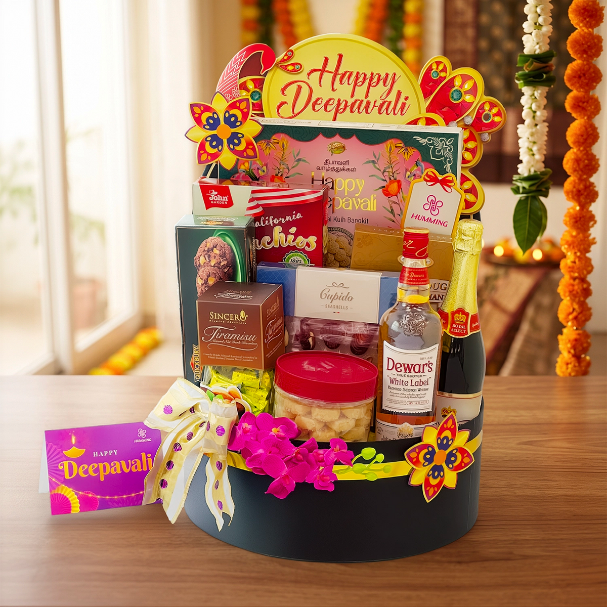 A festive Deepavali gift basket arranged on a wooden table, filled with assorted snacks, chocolates, a bottle of Dewars White Label liquor, a dark bottle, a pink gift box, and a small card. The basket is decorated with a large 'Happy Deepavali' sign, colorful flower ornaments, and a white ribbon with pink polka dots.